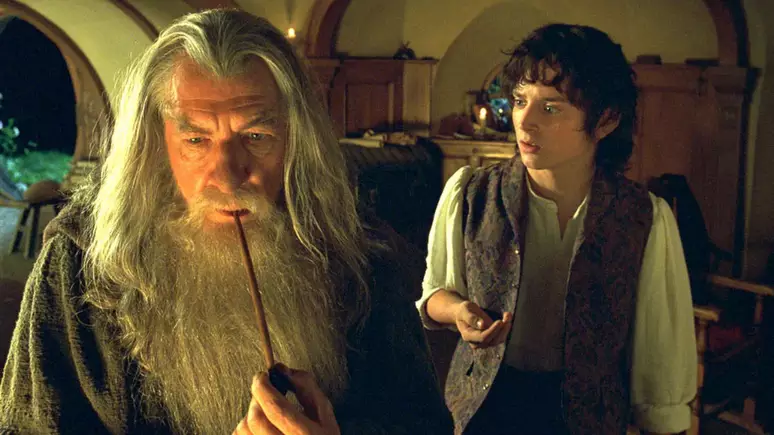 Gandalf fumando cachimbo e olhando para baixo enquanto Frodo olha para ele espantado